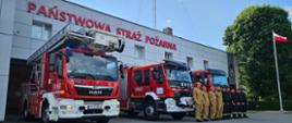 Na zdjęciu widoczna jest grupa strażaków stojących w równym szeregu przed budynkiem jednostki „Państwowa Straż Pożarna”. Po lewej stronie kadru zaparkowane są trzy czerwone samochody strażackie – jeden z podnośnikiem hydraulicznym (oznaczenie 541z54), drugi z oznaczeniem 541z21, a trzeci częściowo widoczny.
Strażacy ubrani są w dwa typy mundurów – beżowe i czarne, ustawieni na tle budynku z dużym czerwonym napisem „PAŃSTWOWA STRAŻ POŻARNA”. Po prawej stronie kadru widać maszt z powiewającą biało-czerwoną flagą Polski oraz duże drzewo. Niebo jest częściowo zachmurzone, a całość tworzy oficjalny, reprezentacyjny charakter zdjęcia.
