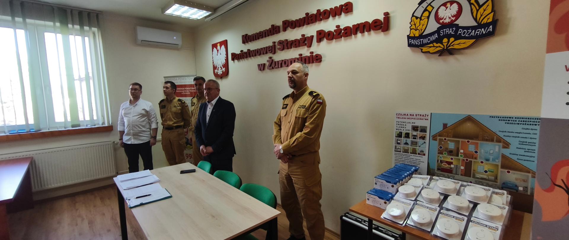 Zdjęcie przedstawia oficjalne spotkanie w Komendzie Powiatowej Państwowej Straży Pożarnej w Żurominie, zorganizowane w ramach Ogólnopolskiego Turnieju Wiedzy Pożarniczej „Młodzież Zapobiega Pożarom”. Na pierwszym planie widoczna jest ekspozycja nowoczesnych czujek dymu i tlenku węgla oraz materiałów edukacyjnych, które mają promować bezpieczeństwo i świadomość przeciwpożarową wśród uczestników.