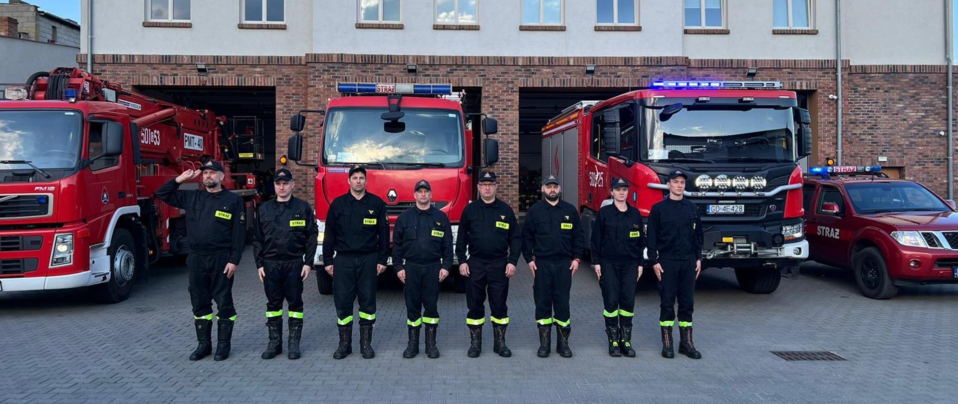 Zdjęcie przedstawia moment oddania hołdu minutą ciszy śp. druhowi Marcinowi Raczykowi, Zmiana I z Jednostki Ratowniczo Gaśniczej ustawiona w szeregu. Pierwszy z lewej dowódca zmiany asp. Adam Liss oddaje honor a reszta osób stoi na baczność. Za funkcjonariuszami ustawione są pojazdy gaśnicze. Strażacy są ubrani w stroje koszarowe, czapki na głowie i czarne buty.