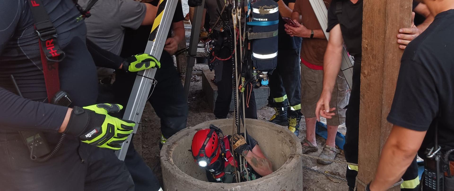 Widok z boku. Na pierwszym planie strażacy, którzy przy życiu trójnogu ratowniczego rozstawionego nad studnią opuszczają ratownika w głąb studni.