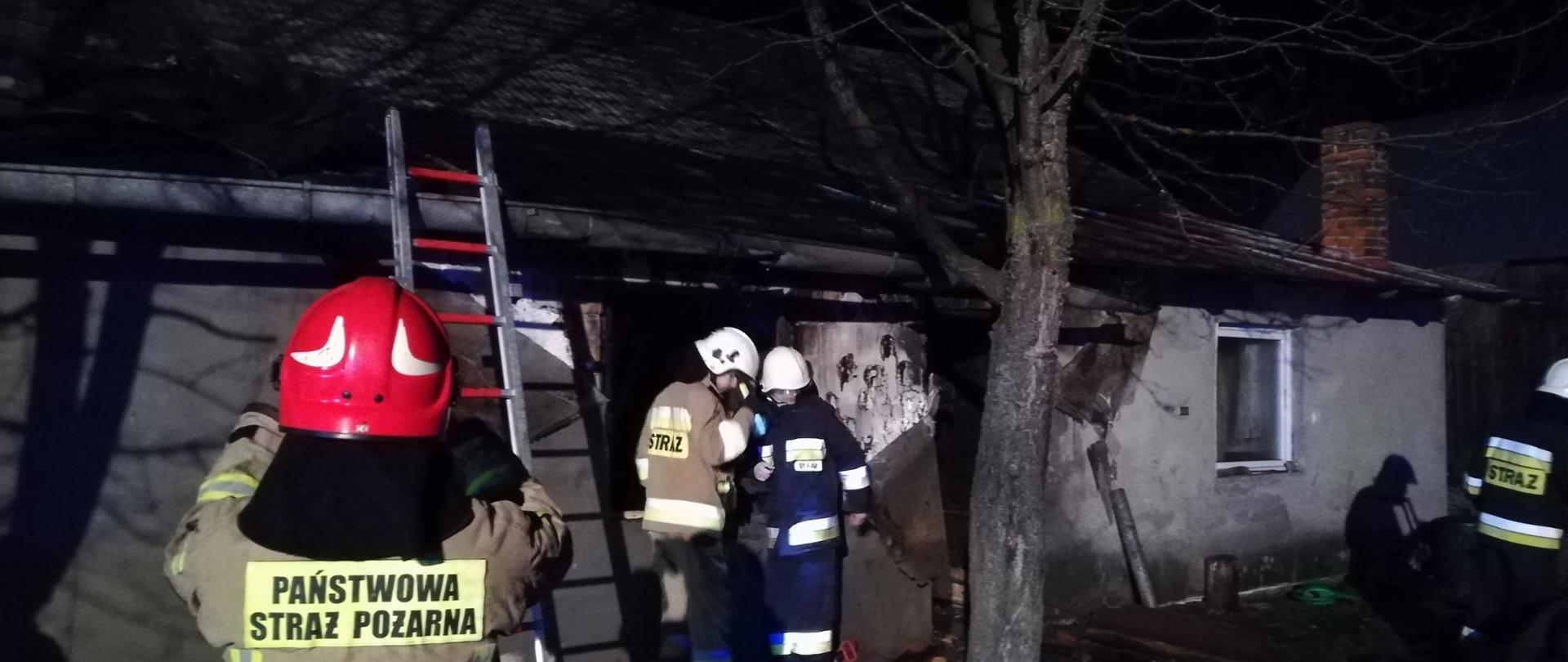 Zdjęcie przedstawia strażaka Państwowej Straży Pożarnej na tle spalonego budynku mieszkalnego. Na drugim planie widoczni strażacy ochotnicy