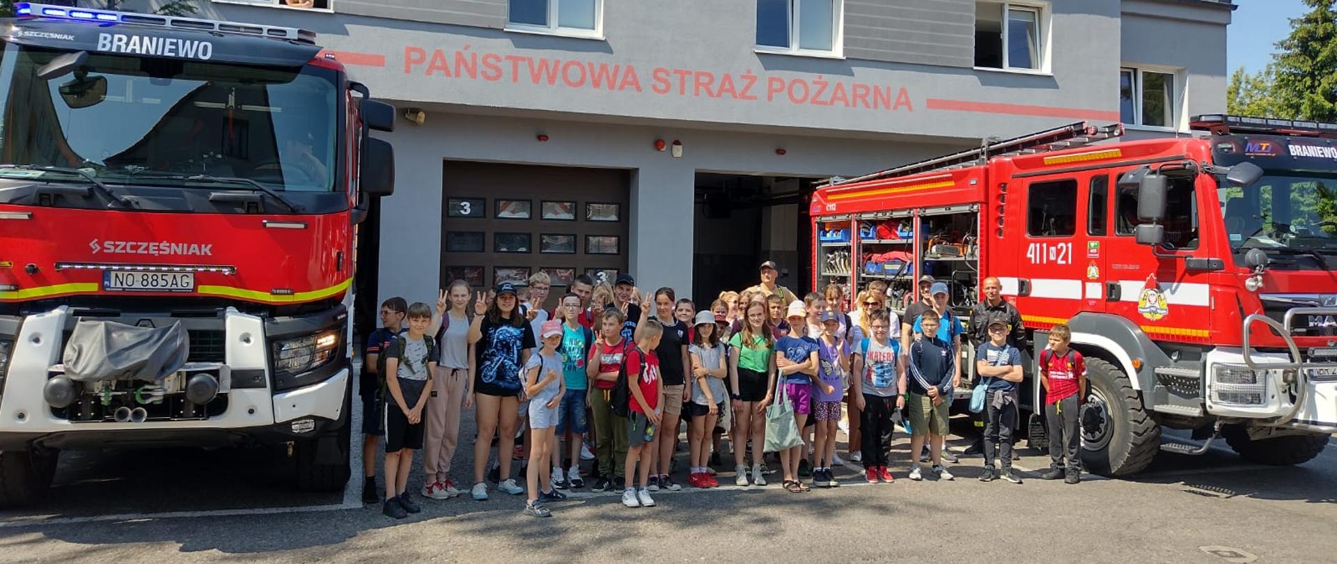 Na zdjęciu grupa dzieci, stoją pomiędzy dwoma samochodami strażackimi. W tle budynek strażnicy.