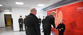 Strażak w mundurze wyjściowym gratuluje chłopcu podając rękę. Po prawej czerwony baner PSP. W tle inni uczestnicy turnieju. Na środku stoi jeszcze drugi strażak w mundurze.