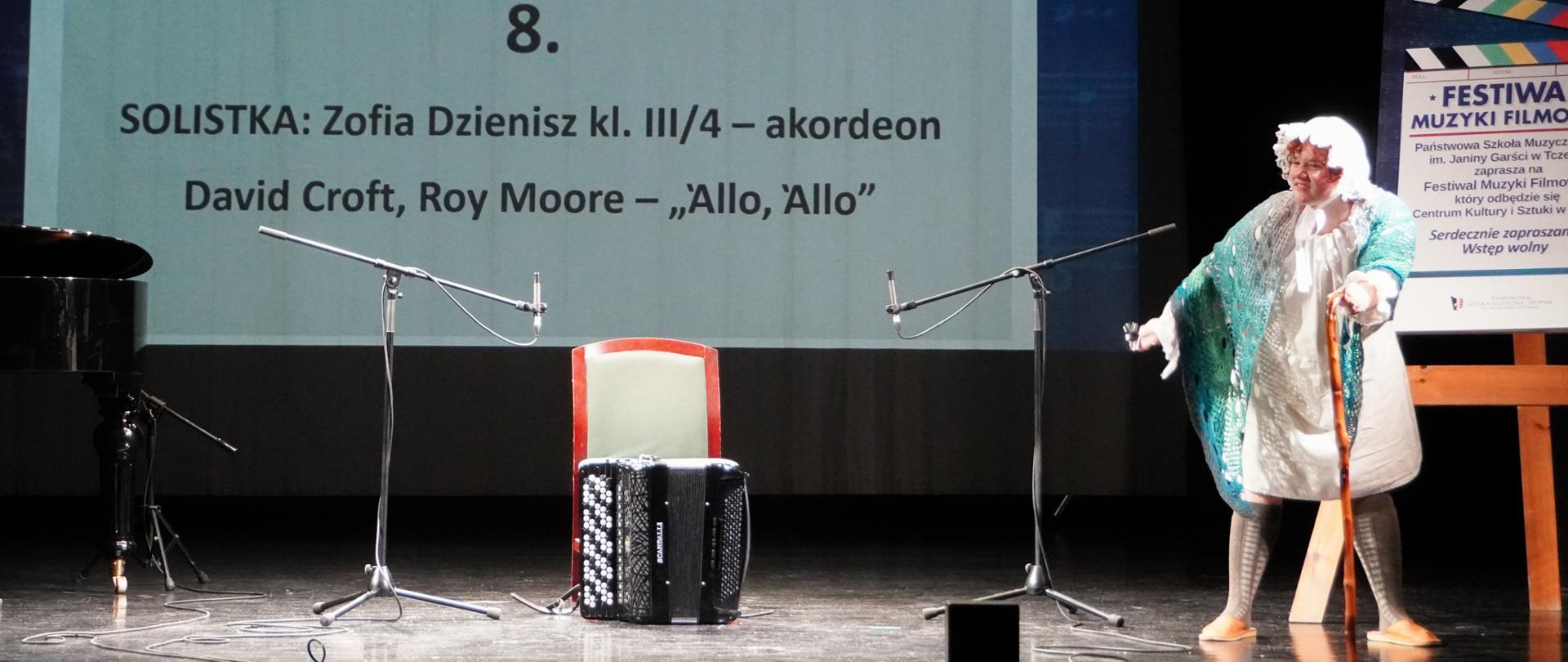 Na scenie tczewskiego CKiS Zofia Dzienisz odgrywa scenkę w przebraniu tuż przed konkursowym wykonaniem. W centrum kadru stoi akordeon, w tle plansza z informacją dotyczącą jej repertuaru.