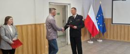 Na zdjęciu widać moment wręczenia wyróżnienia podczas uroczystości w sali o jasnych ścianach i drewnianej boazerii. Mężczyzna w mundurze strażackim podaje dłoń mężczyźnie w koszuli i jeansach, jednocześnie trzymając w drugiej ręce czerwoną teczkę z godłem. Obok stoi kobieta w jasnej marynarce, również trzymająca czerwoną teczkę. W tle widoczne są flagi Polski i Unii Europejskiej oraz ekran projekcyjny.