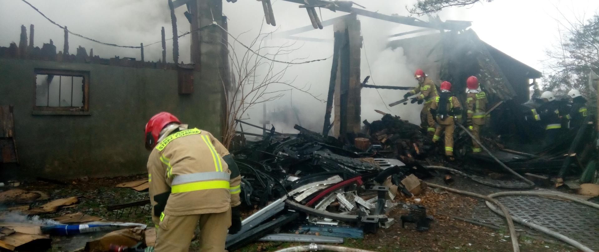 Zdjęcie przedstawia strażaków dogaszających pożar budynku gospodarczego. Na budynku nie ma już dachu - widać resztki spalonej więźby. Panuje lekkie zadymienie. Na ziemi widać węże pożarnicze.