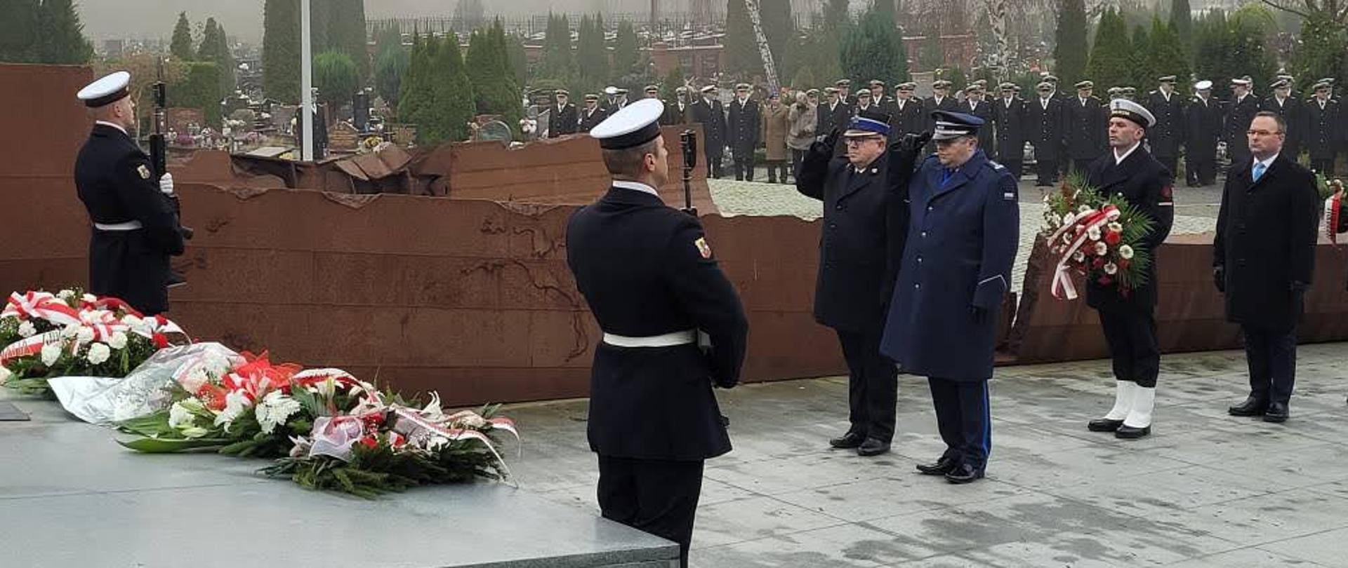 Żołnierze z karabinami stoją przy grobie na którym leżą wiązanki policjant i strażak stoją przed grobem oddając honor za nimi stoi żołnierz z wiązanką oraz mężczyzna.