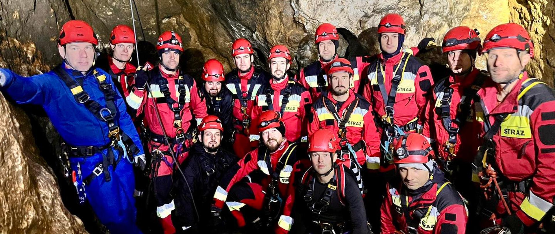 Na zdjęciu widzimy grupę kilkunastu osób wewnątrz jaskini. Wszyscy mają na sobie kaski ochronne w kolorze czerwonym z czołówkami (lampkami czołowymi) oraz uprzęże asekuracyjne. Większość ubrana jest w czerwone kombinezony z żółtymi elementami odblaskowymi i naszywkami z napisem „STRAŻ”. Po lewej stronie stoi mężczyzna w niebieskim kombinezonie, również wyposażony w kask i sprzęt asekuracyjny. Tło stanowią skalne ściany oświetlone sztucznym światłem. Grupa stoi i kuca blisko siebie, patrząc w stronę aparatu.