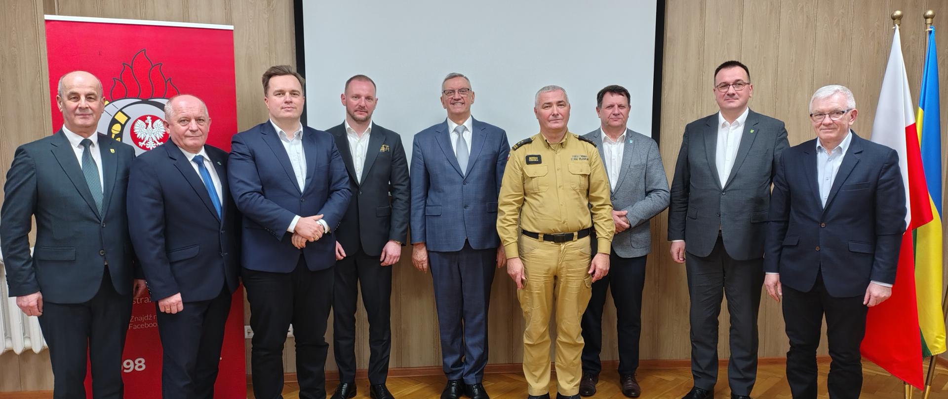 Zdjęcie grupowe wykonane w sali konferencyjnej po naradzie służbowej rocznej podsumowującej działalność ratowniczą w 2025 roku, przedstawia dziewięciu mężczyzn stojących w rzędzie na tle pustego ekranu projekcyjnego oraz drewnianych paneli ściennych. Centralną postacią jest Komendant w piaskowym mundurze służbowym Państwowej Straży Pożarnej, którego otaczają mężczyźni ubrani w garnitury - władze jednostek samorządu terytorialnego. Po lewej stronie grupy widoczny jest fragment czerwonego banera z logotypem straży, natomiast po prawej stronie stoją flagi: Polski, Ostrołęki. Uczestnicy stoją na jasnym parkiecie ułożonym w jodełkę. Zdjęcie pamiątkowe.