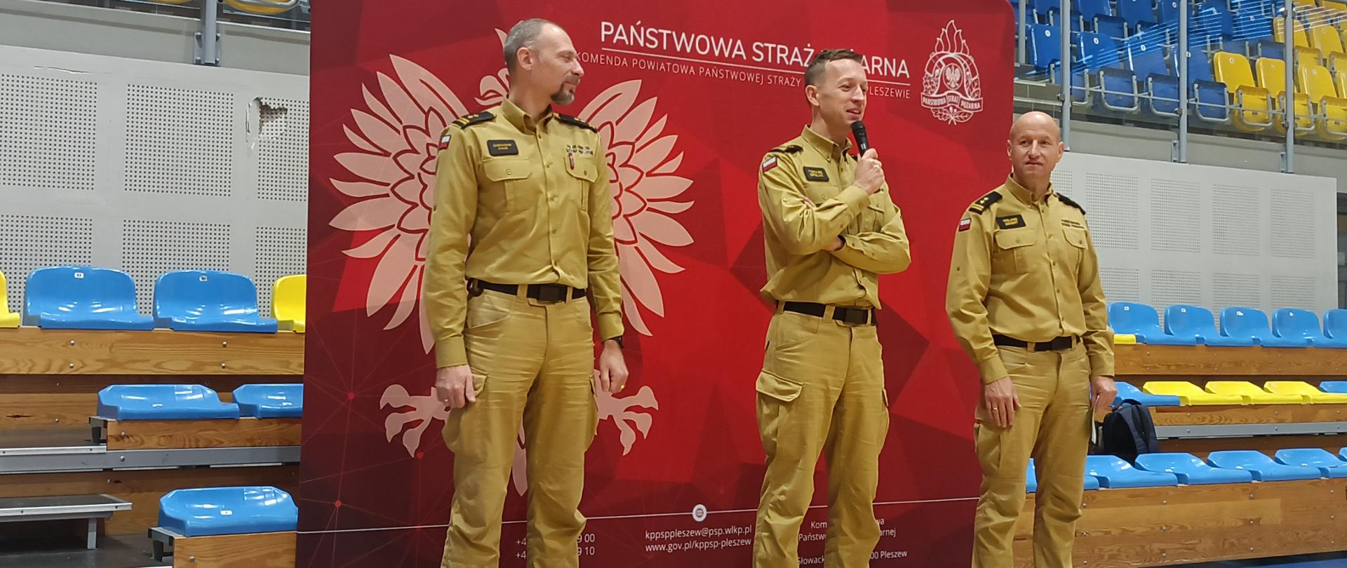 zastępca komendanta wojewódzkiego psp otwiera mistrzostwa piłki nożnej, obok stoi komendant powiatowy z pleszewa oraz zastępca naczelnika ośrodka szkolenia KW PSP w ponznaniu, wszyscy w w piaskowych mundurach, w tle siedzenia w hali sportowej oraz czerwony baner z orłem