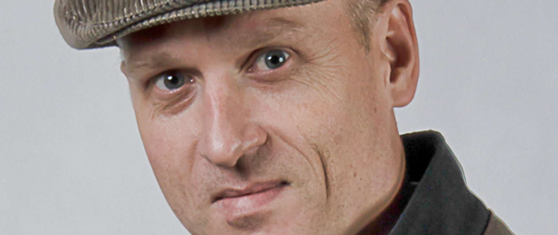 Na zdjęciu widzimy portret Marka Króla-Józagi dyrektora szkoły. Autorką fotografii jest Magdalena Ciszewska-Rząsa. Dyrektor ma na głowie założony sztruksowy kaszkiet w kolorze beżowym. Postać zwrócona jest w kierunku widza i jest lekko uśmiechnięta. Marek Król-Józaga ma założoną białą koszulę w delikatną błękitną kratkę z krawatem w pastelowe paski, ma również ubraną granatową kurtkę. Tło jest białe