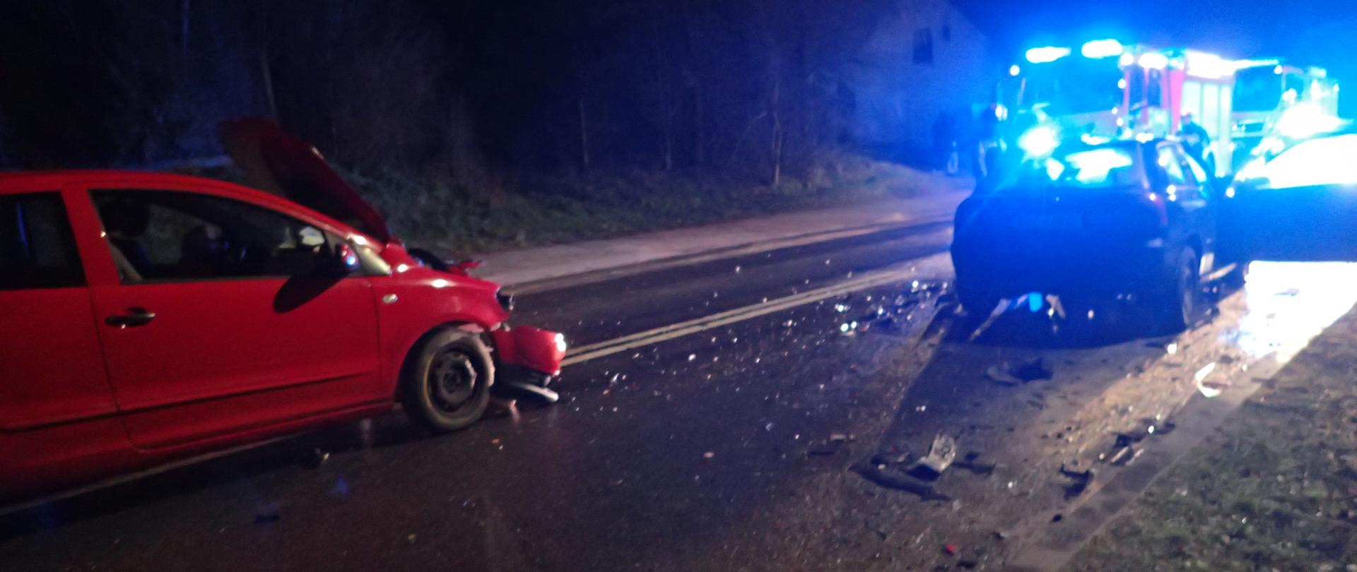 Dwa pojazdy po zderzeniu czołowym znajdujące się na jezdni, pora nocna w tle samochód pożarniczy na sygnałach świetlnych