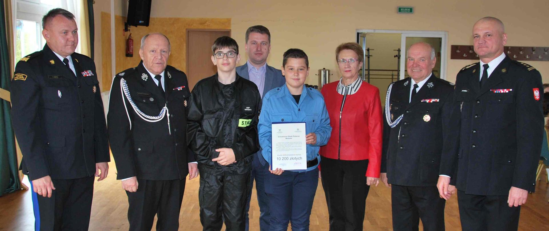 W pomieszczeniu na dużej Sali stoi dwóch strażaków w mundurach PSP, dwóch strażaków w mundurach OSP mężczyzna w garniturze oraz kobieta w czarno-czerwonym stroju. Pomiędzy nimi stoi chłopiec prezentując przed sobą dokument.