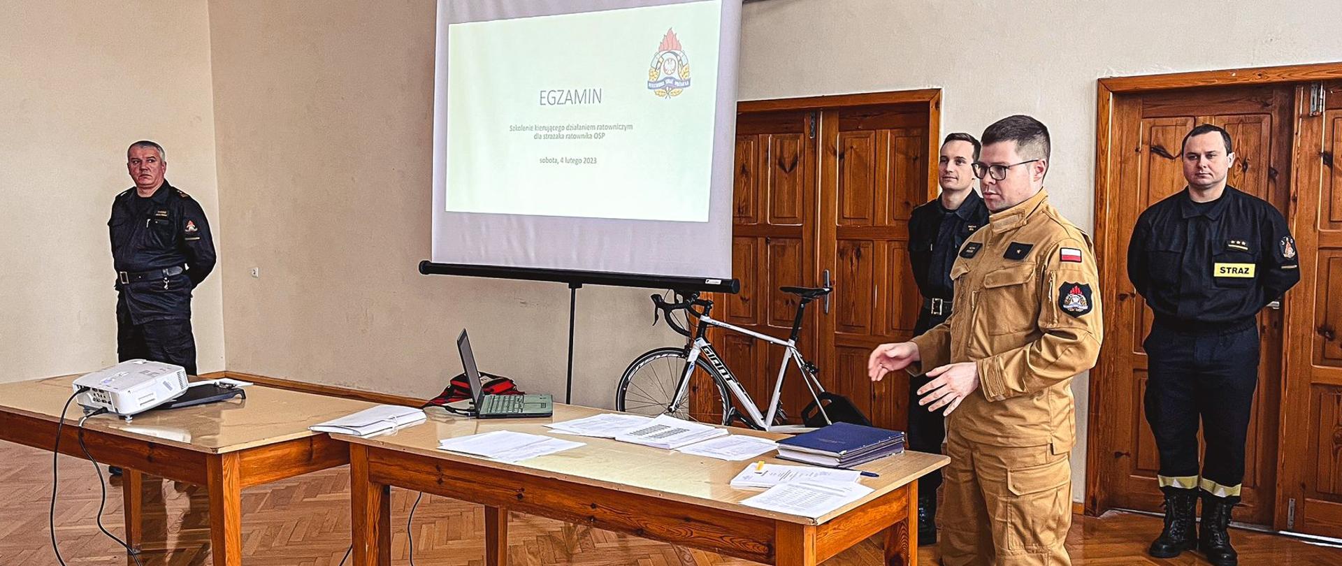 Komisja egzaminacyjna w trakcie omawiania egzaminu. Osoby stoją przy stole, na drugim planie wyposażenie sali gimnastycznej oraz ekran projektora. Zdjęcie wykonane w sali gimnastycznej strażnicy.