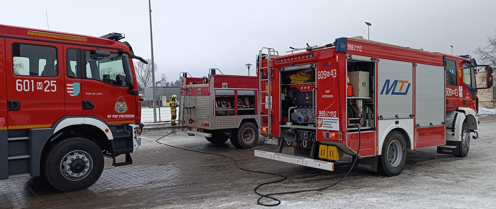Zdjęcie przedstawia trzy czerwone wozy strażackie zaparkowane na zaśnieżonym placu w ciągu dnia. Na pierwszym planie, po lewej stronie, widać przód dużego wozu strażackiego z numerem "601 25" i napisem "KP PSP PRZYSUCZA". Jest to pojazd gaśniczy, typowy dla polskiej straży pożarnej. W środku kadru znajdują się dwa mniejsze pojazdy, również w kolorze czerwonym. Drugi, po prawej stronie, to kolejny wóz strażacki z numerem "609 43" i logo "MTI", a także numerami alarmowymi "998/112". Z tyłu tego pojazdu widać otwarte schowki, odsłaniające sprzęt ratowniczy. Między pojazdami leżą węże i sprzęt, wskazując na trwające lub niedawno zakończone działania. W tle widać zamglony, jasny krajobraz z kilkoma drzewami i budynkami w oddali, co sugeruje zimowy dzień. Na zdjęciu widać również jedną osobę w żółtym ubraniu strażackim, stojącą między dwoma środkowymi pojazdami. Cała scena wskazuje na operację straży pożarnej w trudnych, zimowych warunkach.