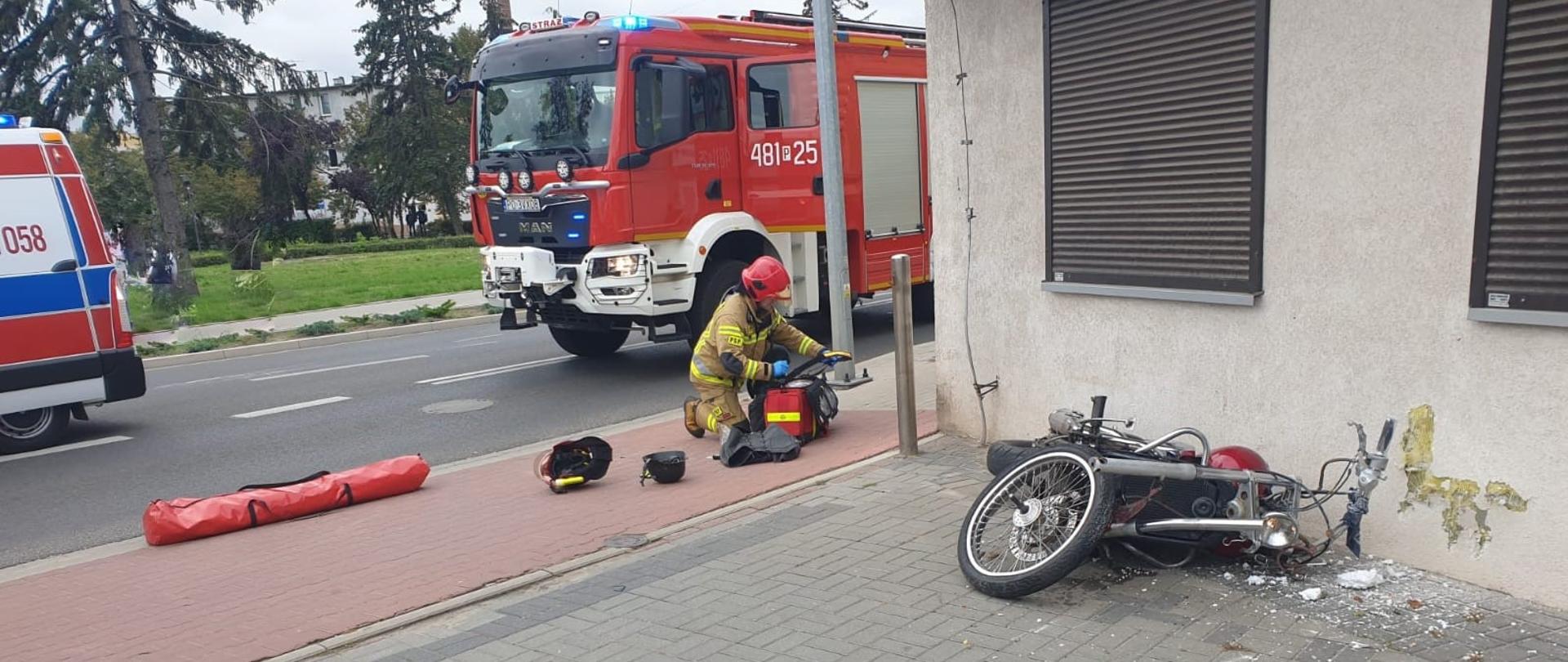 MOTOCYKLEM W ŚCIANĘ BUDYNKU. Motocykl leży obok ściany budynku, widać uszkodzoną elewację. Obok strażak składający sprzęt, w tle samochód strażacki.