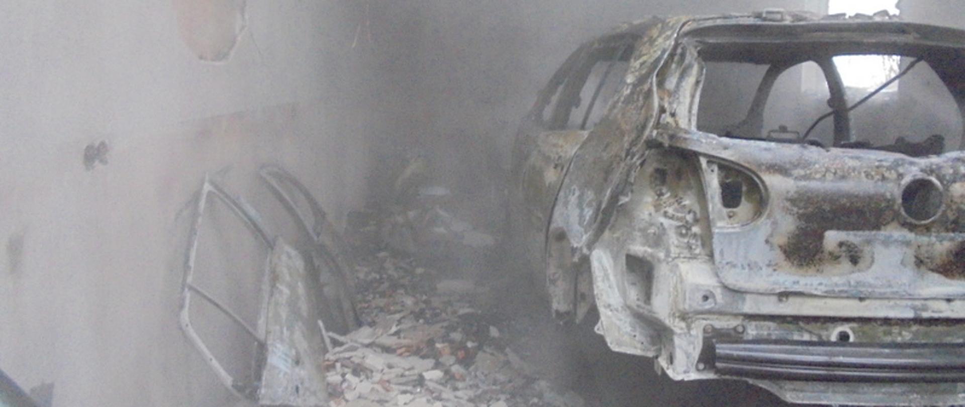  Zdjęcie przedstawia całkowicie spalony samochód osobowy znajdujący się w garażu. Po lewej stronie zdjęcia znajduje się ściana pomieszczenia, z której w wyniku wysokiej temperatury jaka panowała podczas pożaru odpadł tynk. Na ziemi można również zauważyć resztki karoserii oraz dużą ilość odpadniętego tynku. Zdjęcie garażu lekko przysłania unosząca się w pomieszczeniu para wodna powstała w wyniku kontaktu wody z nagrzanymi elementami samochodu.