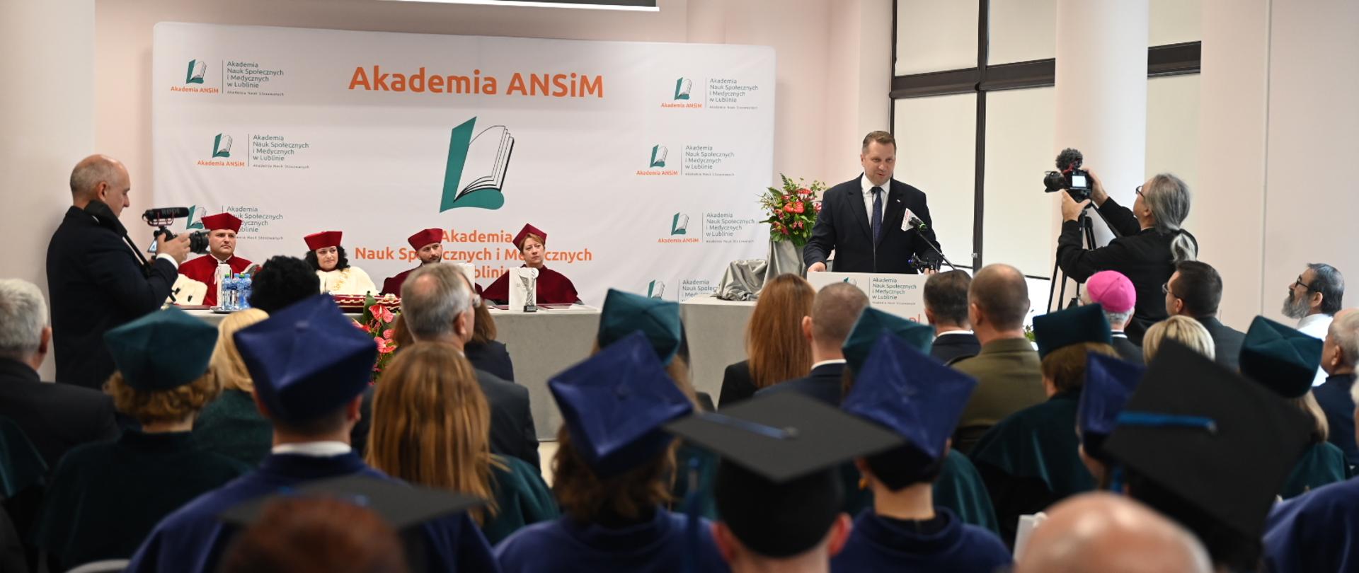 Widok z tyłu na salę, za mównicą stoi minister Czarnek i mówi do mikrofonu, za nim za białym stołem siedzą cztery osoby w czerwonych ceremonialnych strojach, przed nim wypełniona ludźmi sala.