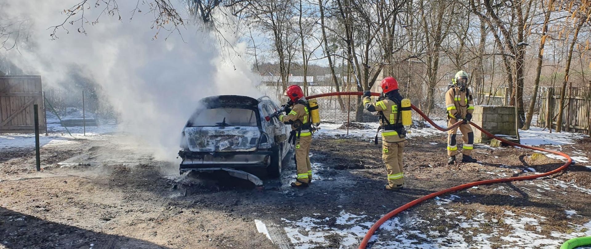 Na zdjęciu widać trzech strażaków w ubraniach specjalnych z założonymi na plecach aparatami ochrony dróg oddechowym. Strażacy gaszą palący się samochód osobowy na prywatnej posesji. W rękach trzymają wąż gaśniczy. Z samochodu wydobywa się gęsty, biały dym. Na ziemi zalega nieznaczna warstwa śniegu.