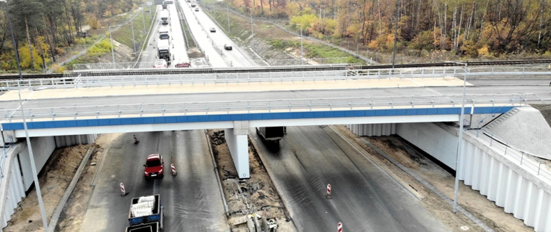 Wiadukt z niebiesko-białymi barierkami nad drogą z ruchem dwukierunkowym, z pracownikami i oznakowaniem robót drogowych; w tle jesienne krajobrazy.