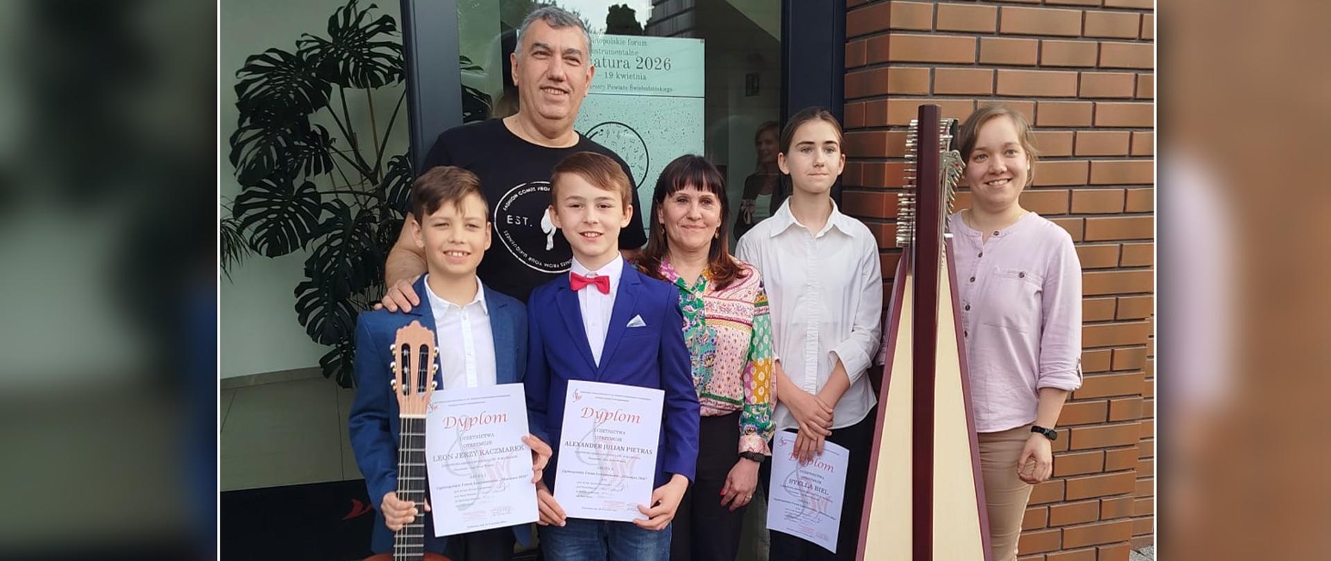 Troje nauczycieli i troje uczniów stoją przed drzwiami szkoły muzycznej. . Uczniowie trzymają w rękach dyplomy.
