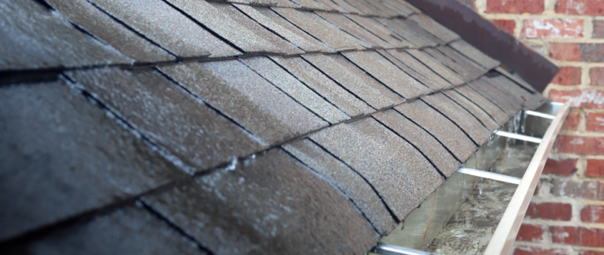 Fragment dachu z ciemnym pokryciem i rynną, na tle ceglanej ściany.