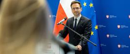 Minister rozwoju i technologii Waldemar Buda podczas konferencji prasowej, minister stoi za mównicą z mikrofonem, za jego plecami flagi Polski i UE oraz niebieski baner z logo MRiT.