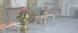 Ćwiczenia w Zakładzie Karnym w Rawiczu. Wnętrze budynku produkcyjnego, w którym ma miejsce pożar. Dwóch strażaków w czerwonych hełmach, w pozycji klęczącej, wyposażeni są w sprzęt ochrony układu oddechowego. Dowódca roty trzyma w rękach prądownicę, symulując gaszenie pożaru. Drugi strażak znajduje się w odległości ok. 2 metrów od dowódcy. Jest odwrócony od niego.