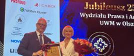 Mężczyzna i Kobieta stoją razem na scenie, Mężczyzna trzyma tabliczkę pamiątkową a kobieta kwiaty 