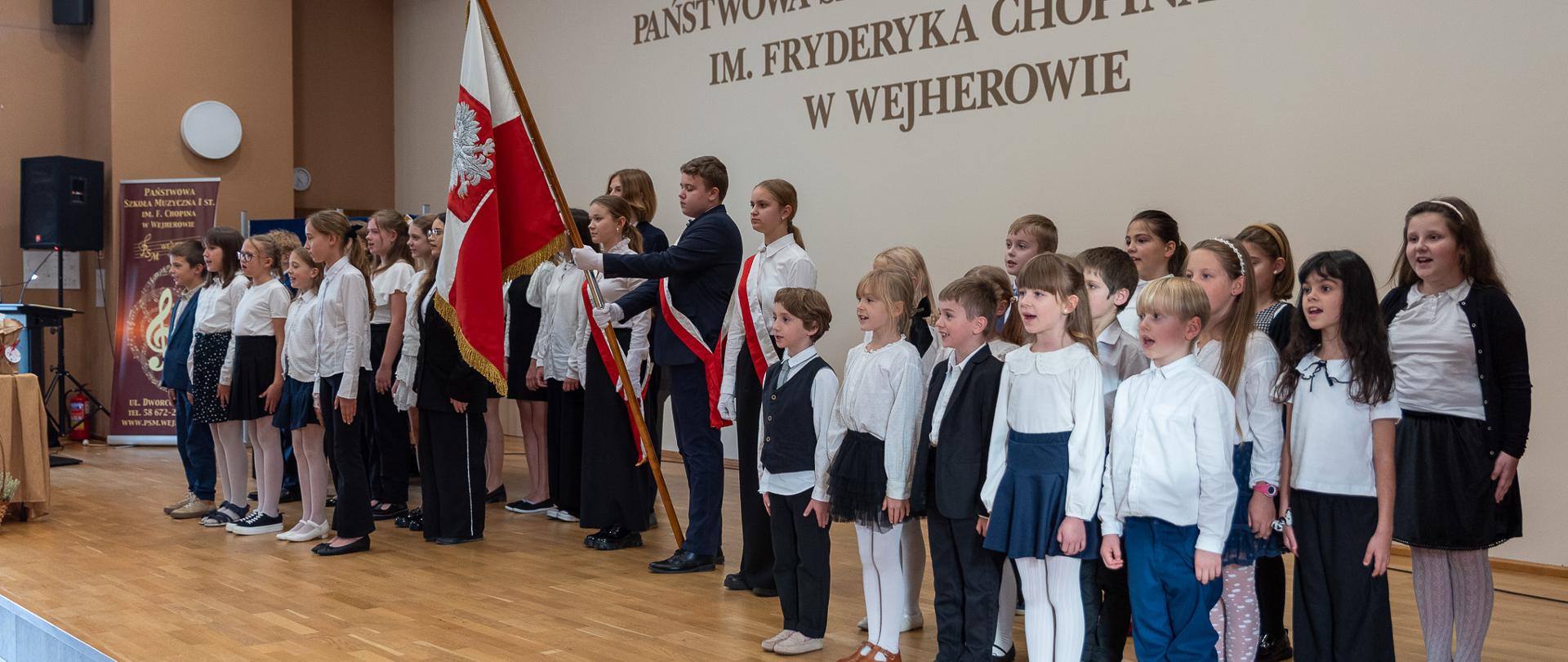 Na scenie sali koncertowej stoją wszyscy uczniowie klas pierwszych i śpiewają Hymn Polski. Po środku poczet sztandarowy wraz ze sztandarem.