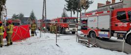 Na zdjęciu widoczne są wozy strażackie. Stoją na zaśnieżonej drodze. z prawej strony widoczni są strażacy w ubranich bojowych oraz hełmach na głowach.