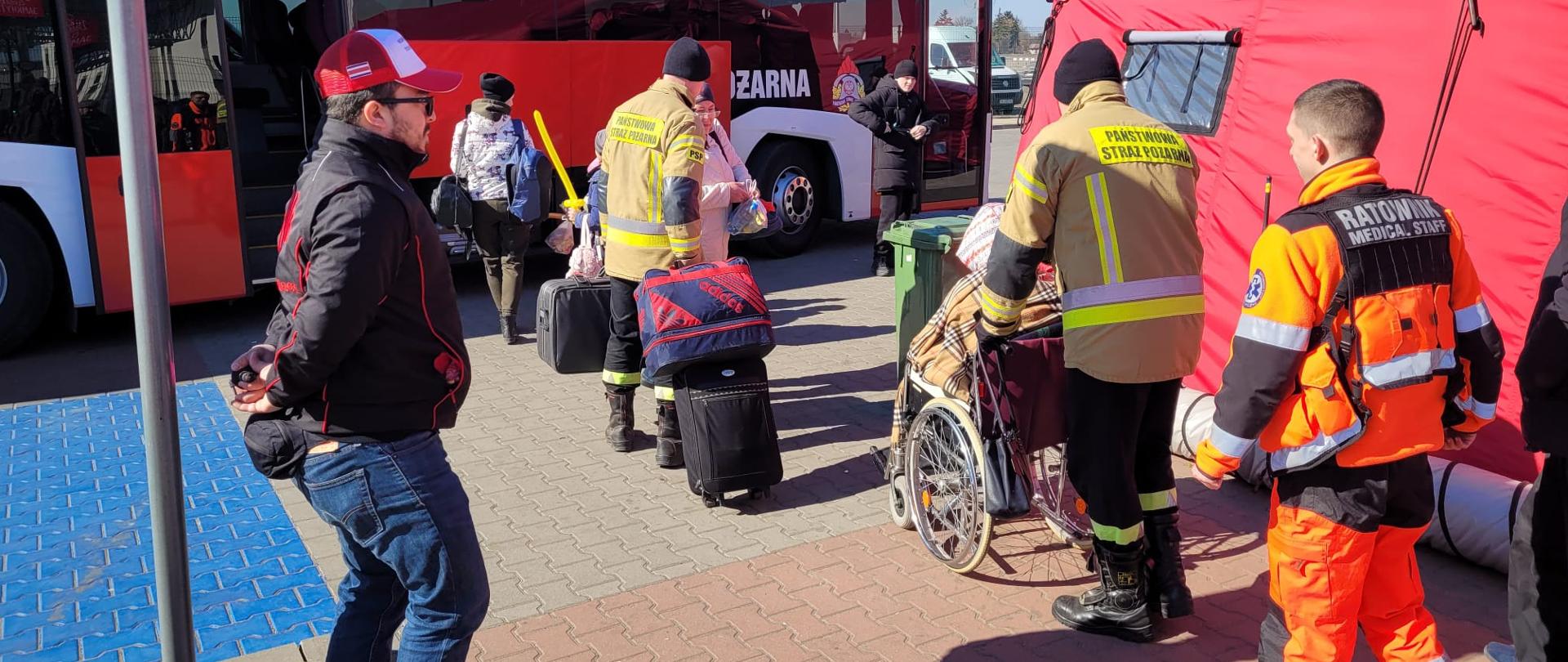 Kadet pomaga osobie na wózku, drugi niesie bagaże. Za nimi ratownik medyczny. W głębi czekający autobus PSP