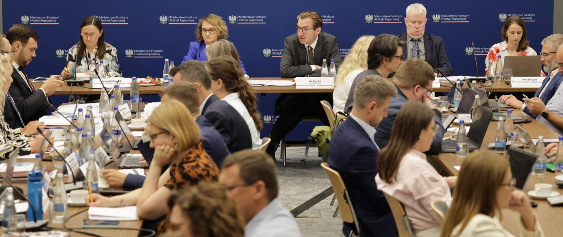 Grupa osób w sali konferencyjnej siedzi przy stołach.