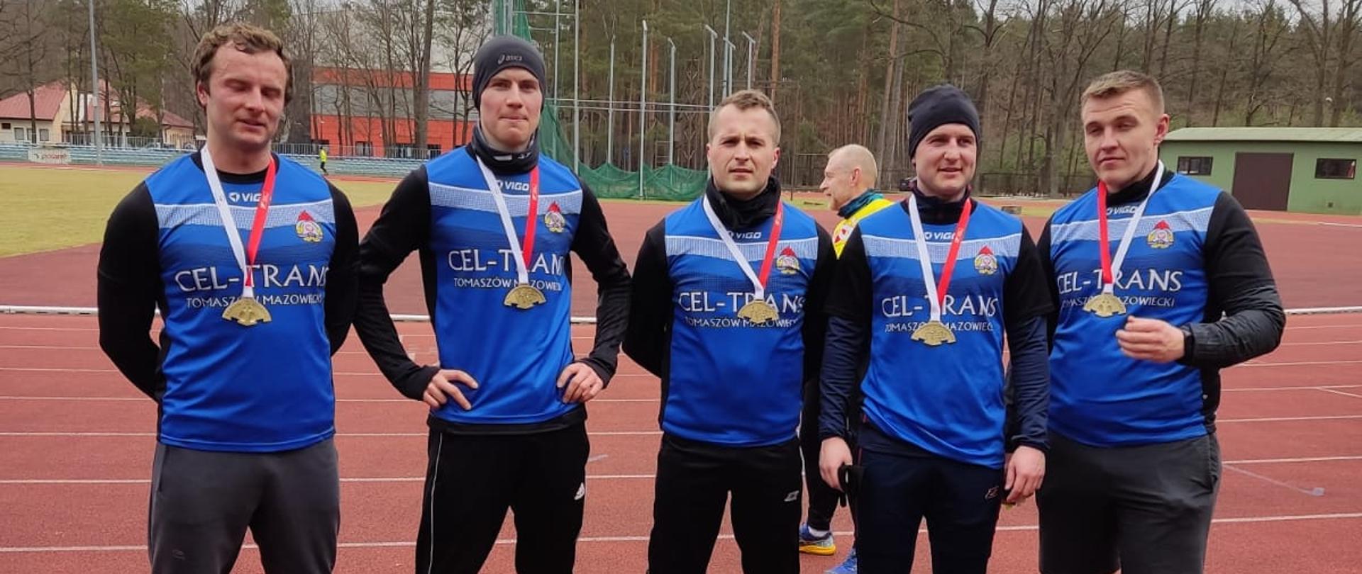 Pięciu mężczyzn w strojach sportowych stoi na bieżni. Dwóch z nich ma na głowie czapki. Wszyscy mają zawieszone na szyj medale 