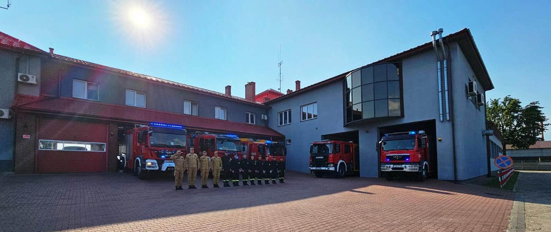 Na zdjęciu widać strażaków KP PSP Skarżysko-Kamienna ubranych w ubrania koszarowe, oddających hołd Powstańcom Warszawskim. Stoją oni przed garażami, są wyprowadzone również samochody bojowe z włączonymi sygnałami świetlnymi i dźwiękowymi. W tle widać budynek Komendy Powiatowej. 