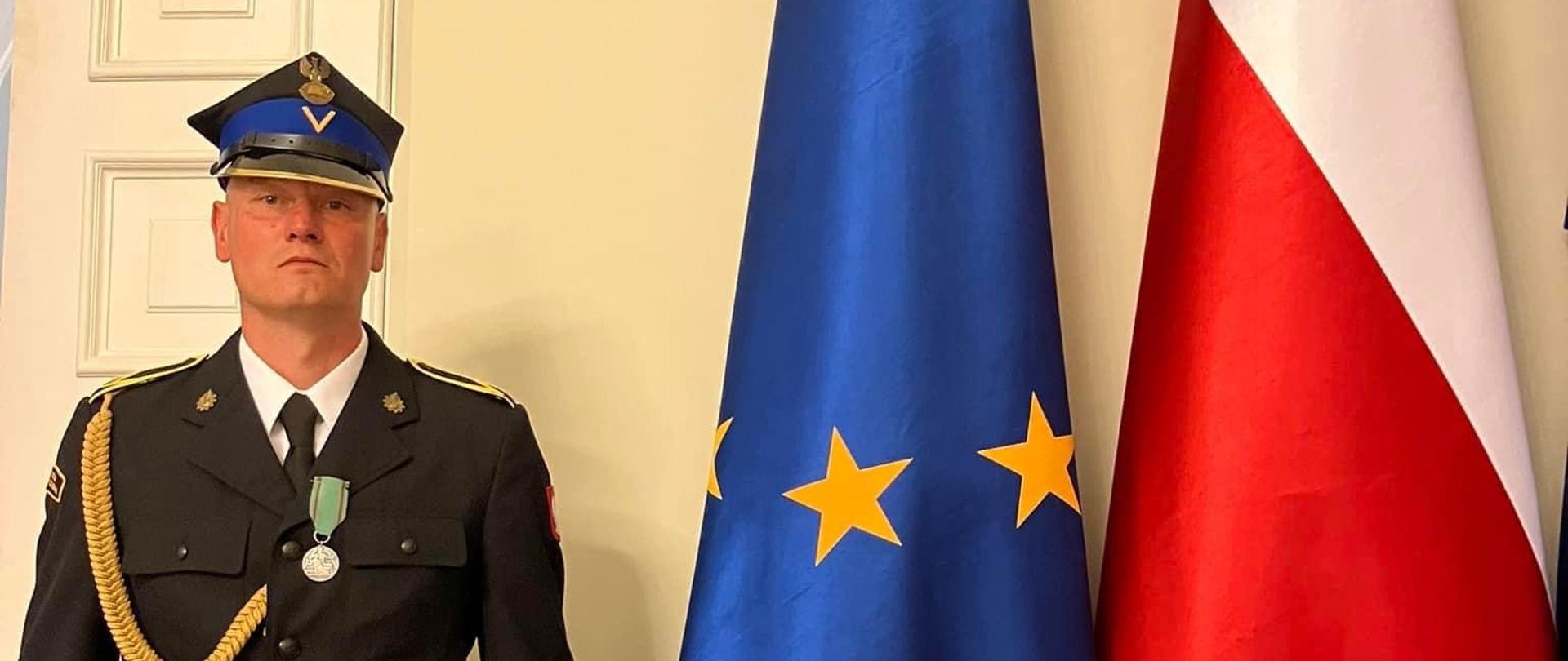 Z lewej ogn. Jacek Sandomierz w granatowym ubraniu specjalnym, z prawej flaga Polski i Unii Europejskiej. Zdjęcie zrobione w korytarzu Pałacu Prezydenckiego, w tle żółta ściana.