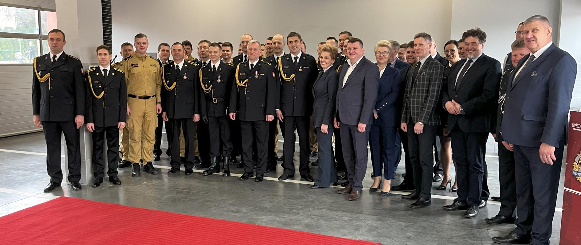 Zdjęcie grupowe uczestników uroczystości stojących w pomieszczeniu garażowym. przed nimi na podłodze widoczny jest duży fragment czerwonego dywanu. 