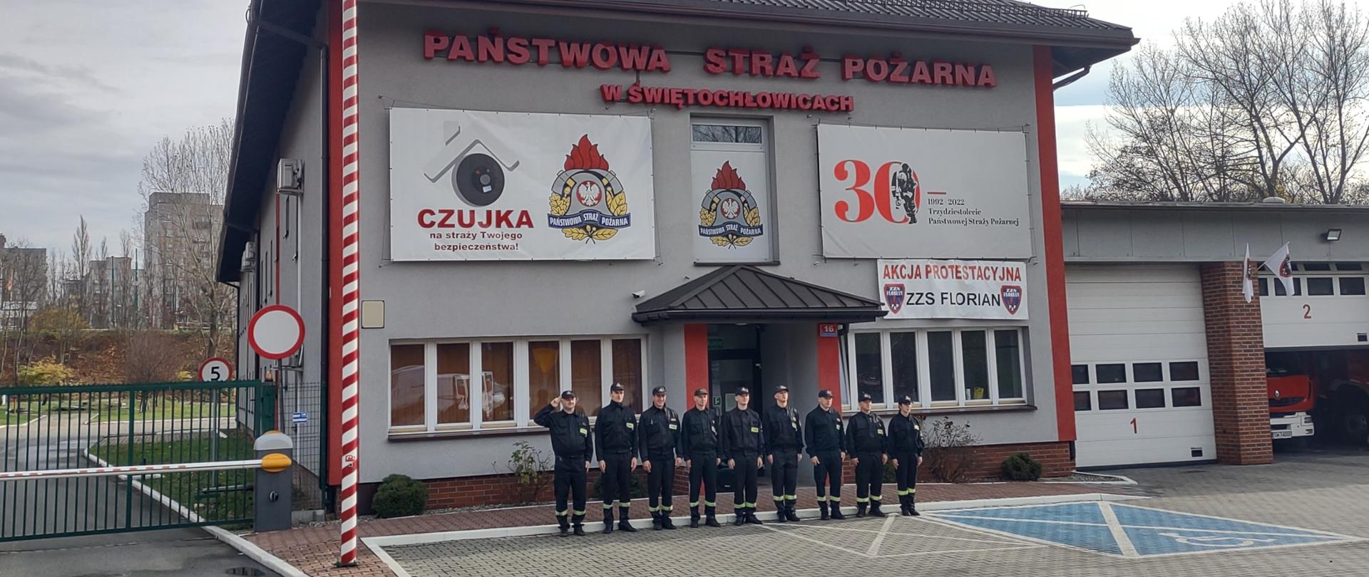 Zdjęcie przedstawia strażaków stojących na baczność śpiewających hymn. W tle budynek komendy.