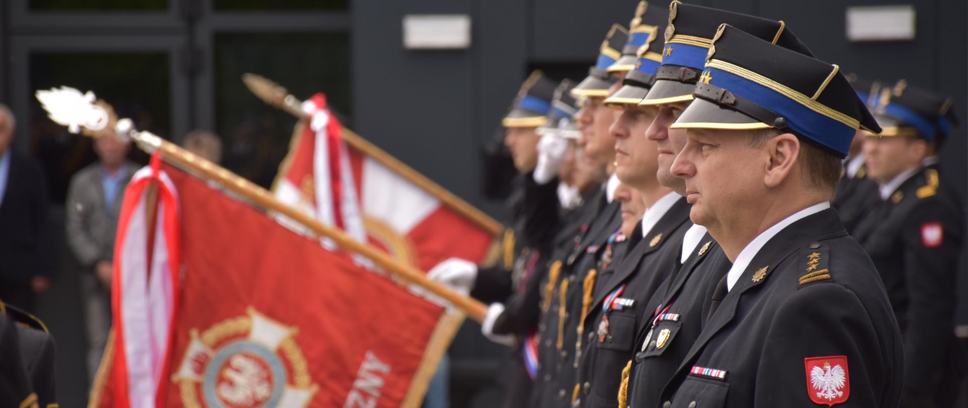 strażacy stojący w szeregu w oddali w szyku pochylone są dwa sztandary, zdjęcie wykonane z boku