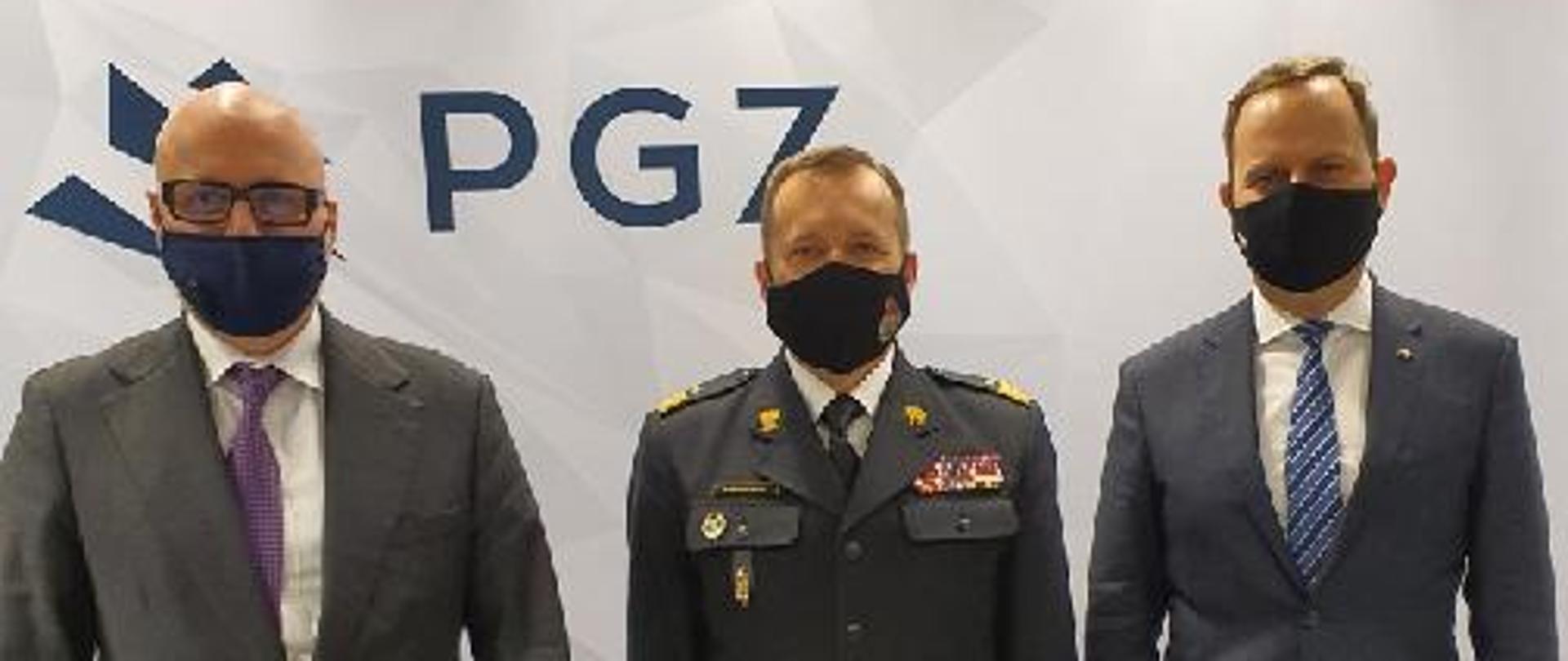 Zastępca komendanta głównego Państwowej Straży Pożarnej w towarzystwie dwóch przedstawicieli przemysłu zbrojeniowego, w tle ściana z logiem Polskiej Grupy Zbrojeniowej