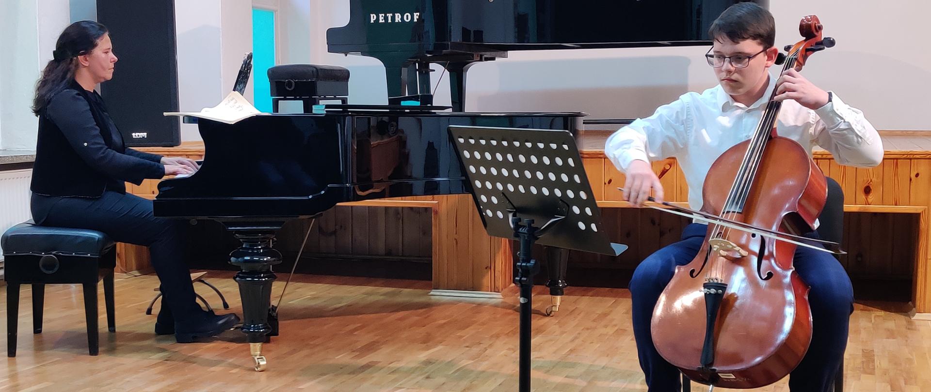 Zdjęcie przedstawiające jednego z uczniów podczas gry na wiolonczeli przy akompaniamencie fortepianowym pani Doroty Straburzyńskiej w trakcie koncertu w auli szkoły w dniu 24 października 2023 roku.