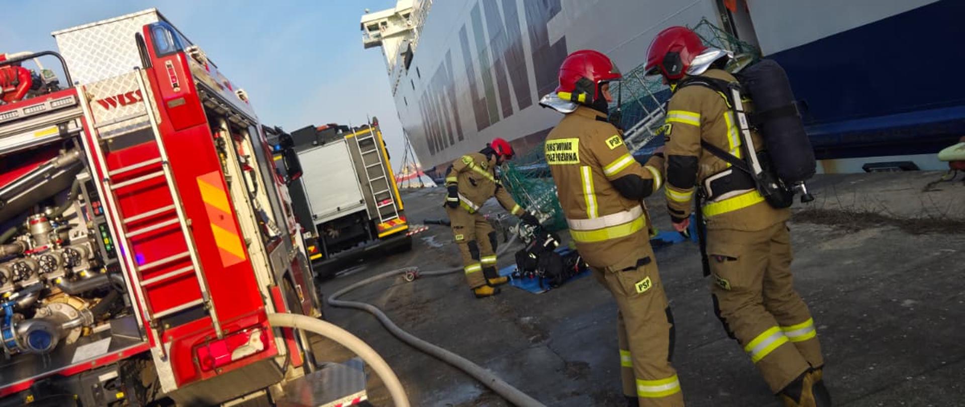 Zdjęcie przedstawia strażaków w trakcie działań przy promie. Po lewej widoczny samochód pożarniczy koloru czerwonego, po prawej dwóch strażaków ubranych w ubranie specjalne koloru piaskowego z czerwonymi hełmami.