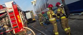 Zdjęcie przedstawia strażaków w trakcie działań przy promie. Po lewej widoczny samochód pożarniczy koloru czerwonego, po prawej dwóch strażaków ubranych w ubranie specjalne koloru piaskowego z czerwonymi hełmami.