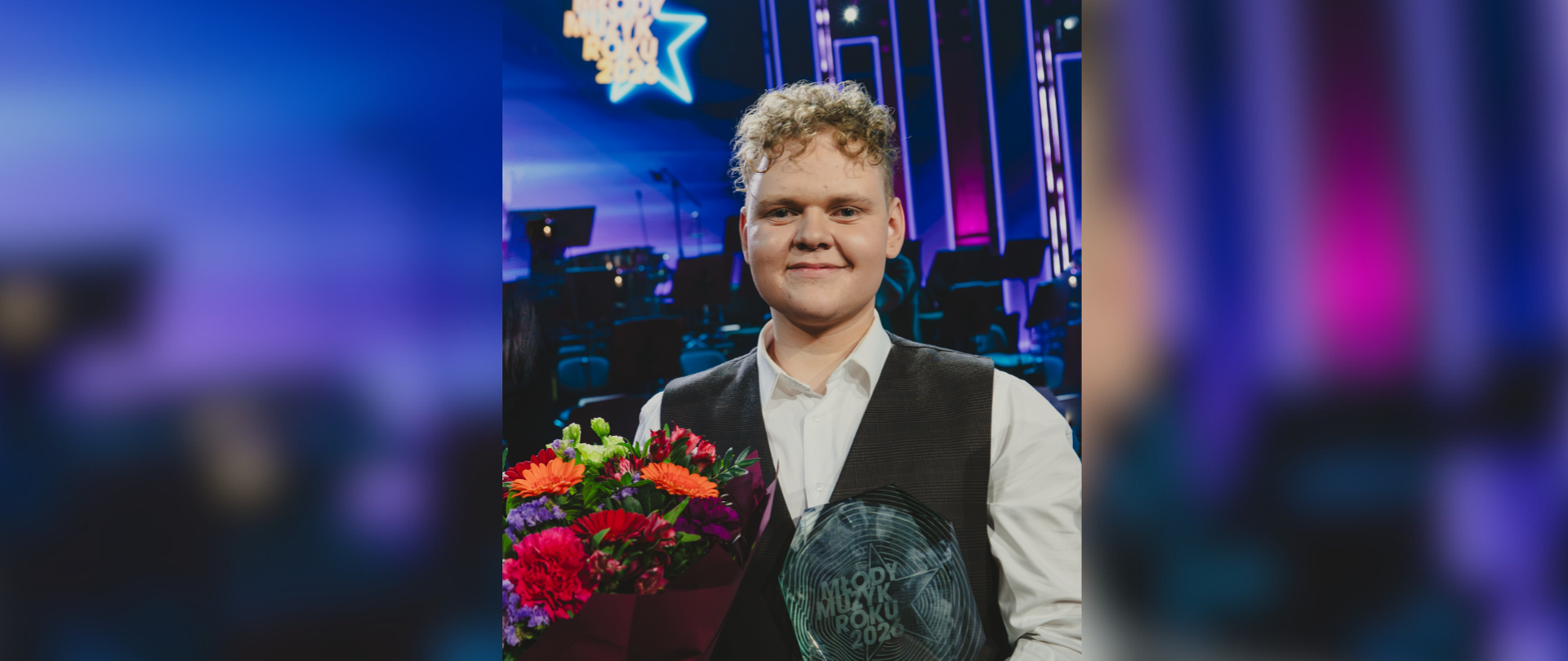  Zapytanie zostało przetworzone
Zdjęcie przedstawia uśmiechniętego młodego mężczyznę, Michała Stochla, zwycięzcę konkursu „Młody Muzyk Roku 2024”. Artysta stoi w blasku scenicznych świateł, ubrany w elegancką białą koszulę i ciemną kamizelkę. W dłoniach trzyma kolorowy bukiet kwiatów oraz szklaną statuetkę konkursową z wygrawerowanym logo wydarzenia. W tle widoczna jest profesjonalna scena muzyczna z instrumentami orkiestrowymi oraz duży, podświetlony neon w kształcie gwiazdy z napisem „Młody Muzyk Roku 2024”, co podkreśla uroczysty charakter chwili i wielki sukces ucznia klasy akordeonu Dariusza Baszaka. Całość kompozycji utrzymana jest w chłodnej, fioletowo-niebieskiej tonacji oświetlenia koncertowego.