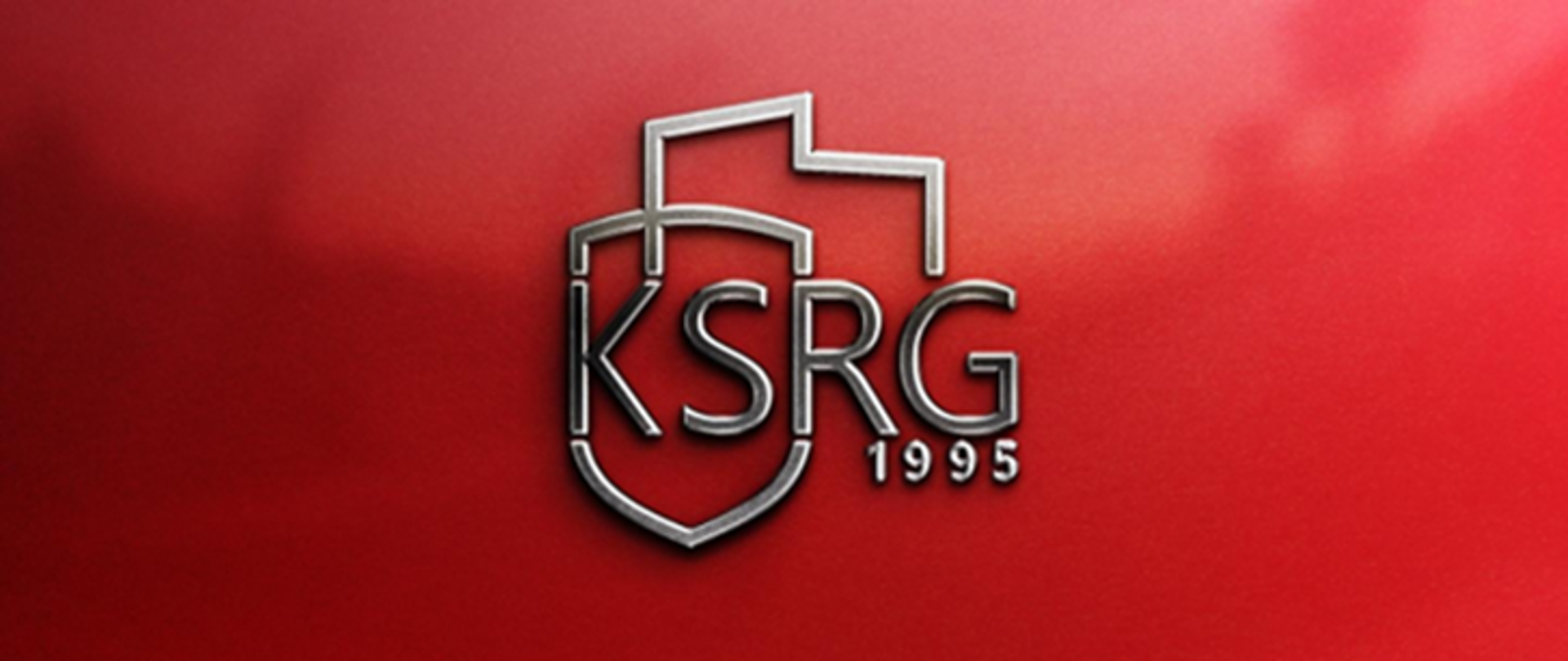 Na ciemnoczerwonym tle widać srebrne, trójwymiarowe logo. Składa się ono z dużej tarczy w kształcie herbu, na której widnieje duży, stylizowany napis „KSRG”. Pod nim, mniejszą czcionką, znajduje się liczba „1995”. Nad tarczą umieszczone są zarysy dwóch budynków, przypominających dach i ściany, a z lewej strony, lekko nachodząc na górną część tarczy, wystaje element przypominający dach lub element architektoniczny. Całość logo ma metaliczny połysk i rzuca delikatny cień, co nadaje mu głębi.