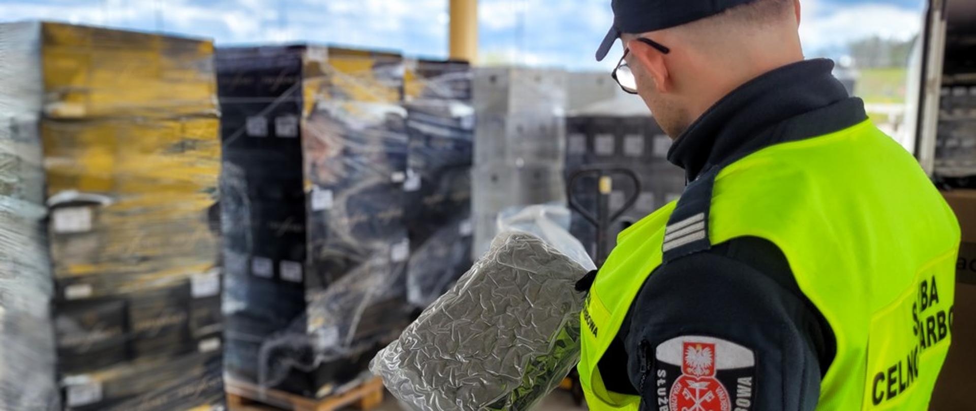 Funkcjonariusz Służby Celno-Skarbowej trzyma przezroczysty, hermetycznie zapakowany pakunek z materiałem roślinnym na tle palet owiniętych folią.