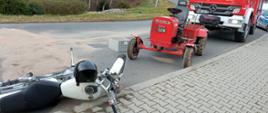 Zdjęcie przedstawia leżący motocykl po zderzeniu z ciągnikiem SAM stojącym obok na jezdni drogi powiatowej. Z prawej strony widoczne pojazdy służb Straży Pożarnej i Policji. W tle zarośla rosnące w pobliżu drogi.