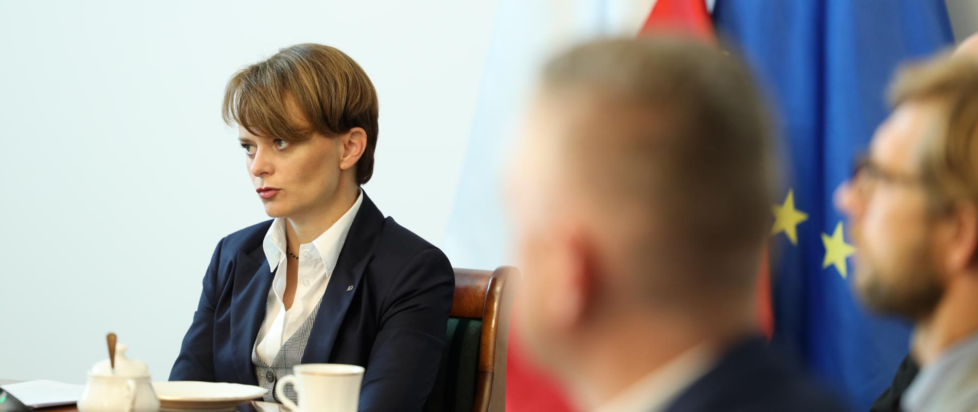 Jadwiga Emilewicz przy stole. Na pierwszym planie niewyraźne profile mężczyzn.