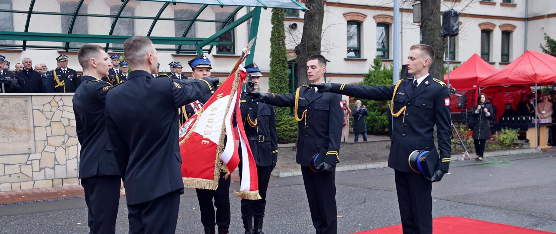 Funkcjonariusze stoją na czerwonym dywanie, składając uroczystą przysięgę przy sztandarze, ręce uniesione w geście ślubowania.
