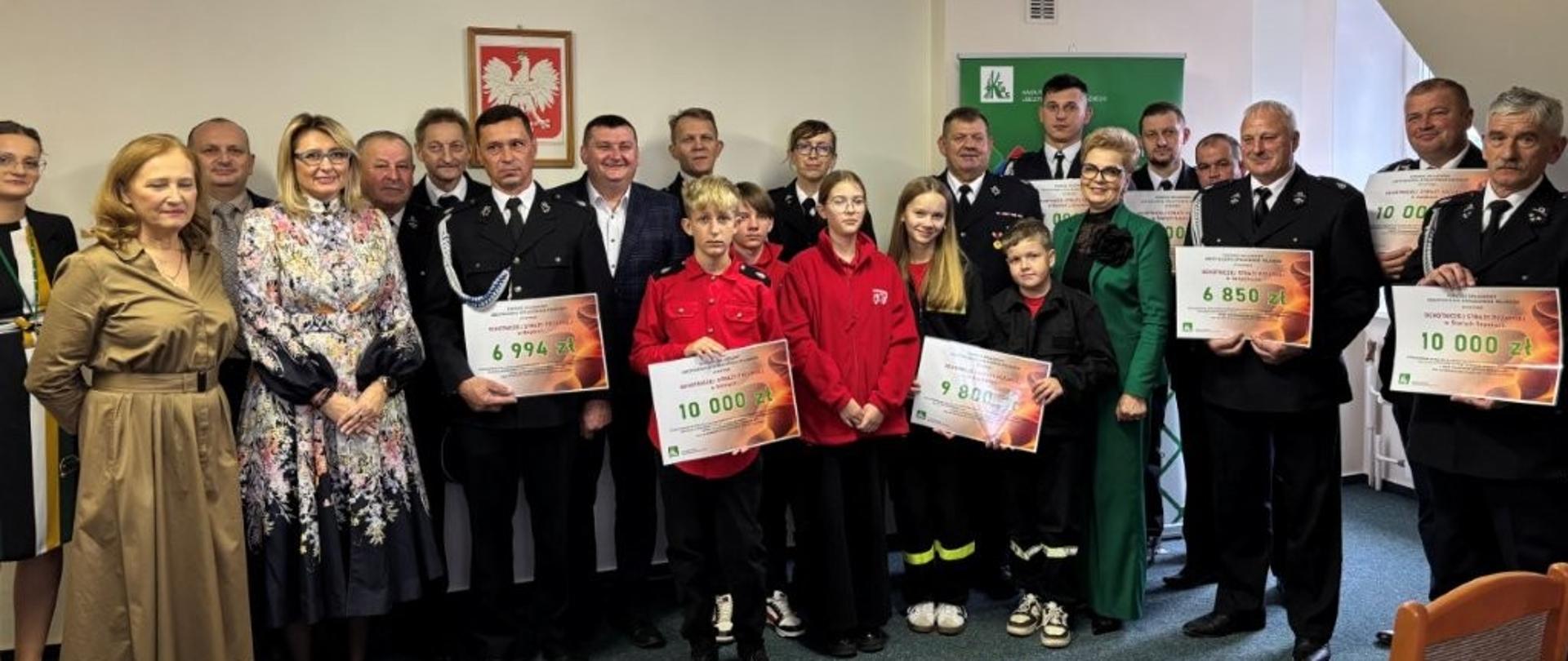 Dwadzieścia trzy osoby mężczyźni i kobiety stoją na tle ściany z godłem polski i banerem KRUS, część z nich w galowych mundurach strażaka trzyma plansze z kwotą otrzymanego wsparcia